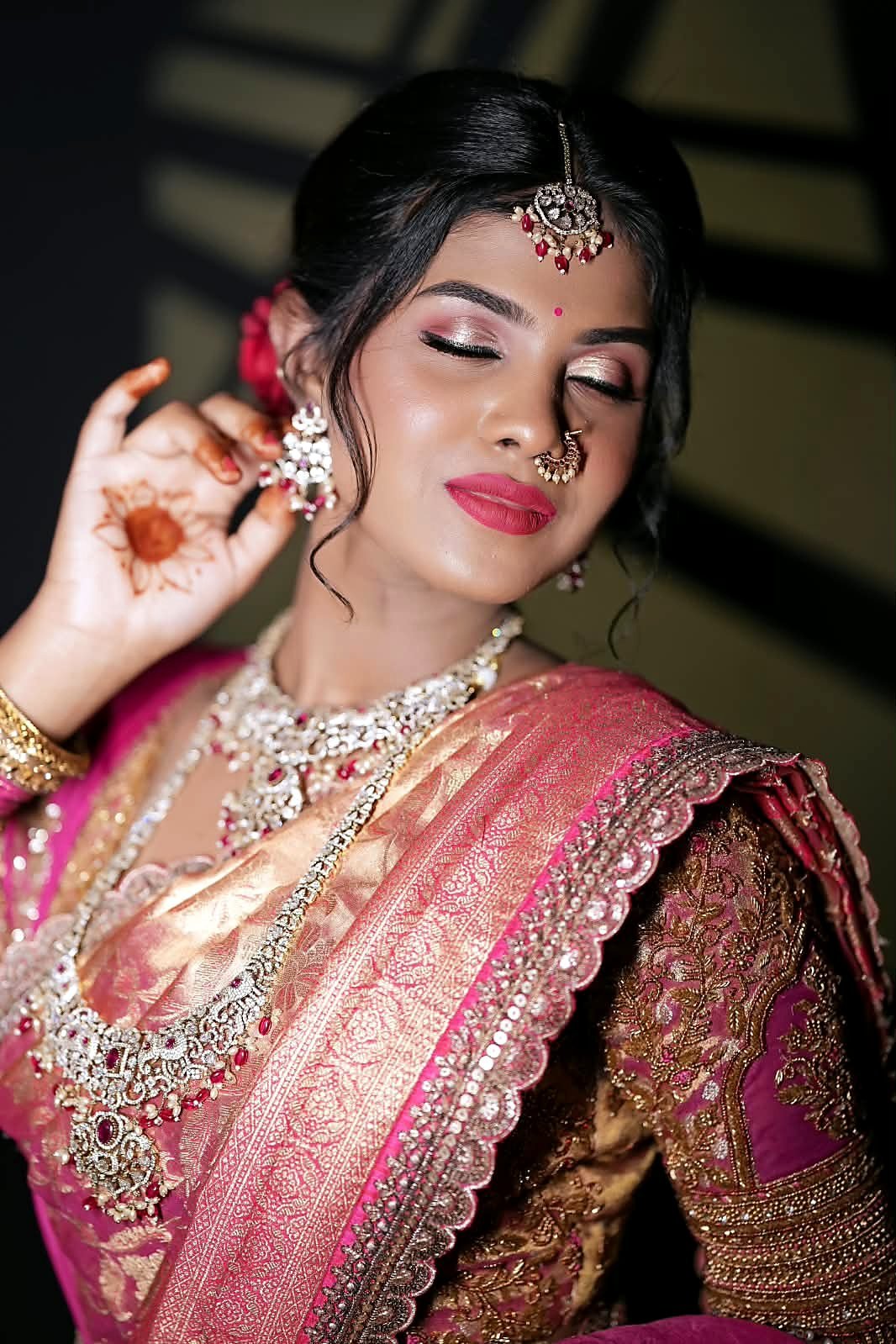 Close-up of bridal eye makeup with shimmering rose-gold lids, defined lashes and pink lips, with henna on the raised hand