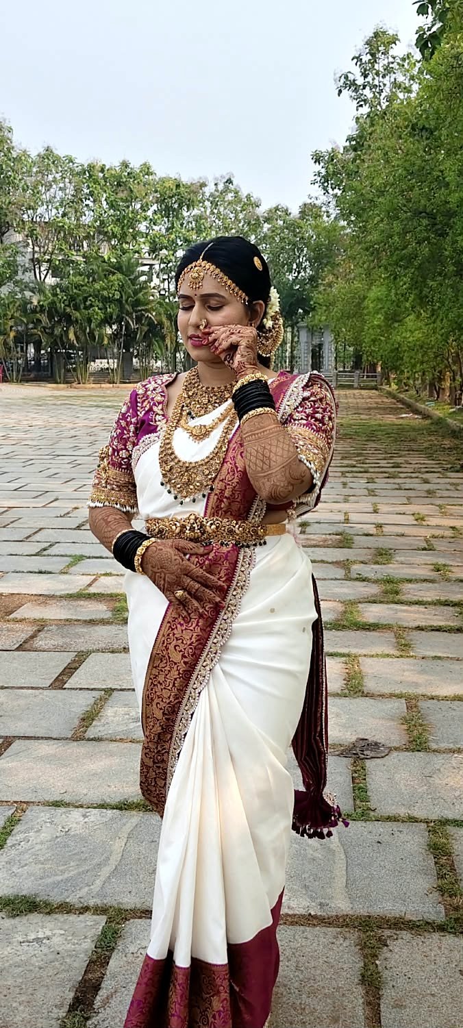 Bride outdoors in an ivory saree with a maroon zari border, an embroidered maroon blouse, gold temple jewellery and bridal mehendi