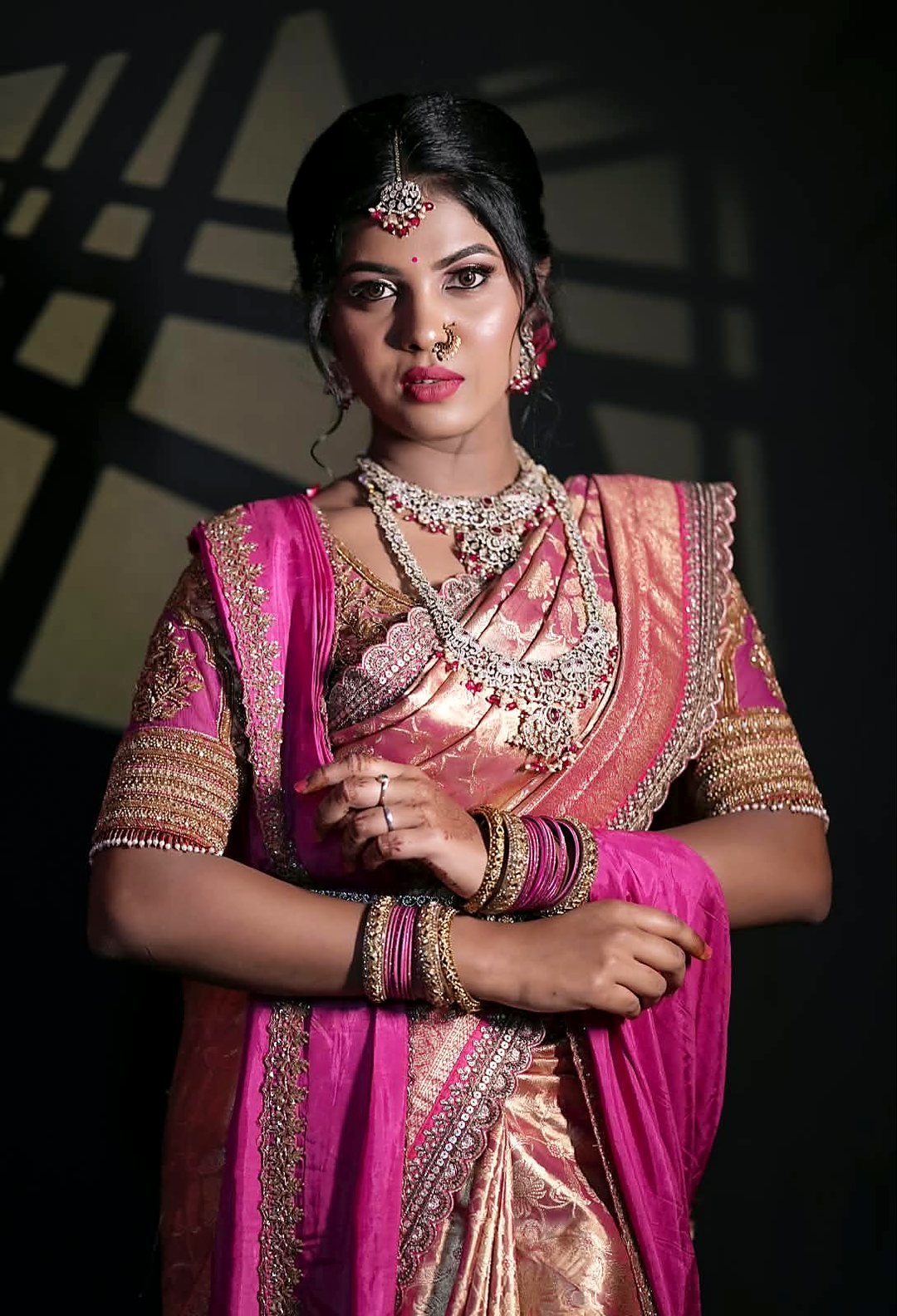 Bride in a pink and gold silk saree with layered diamond-and-ruby necklaces, a maang tikka and soft-glam makeup