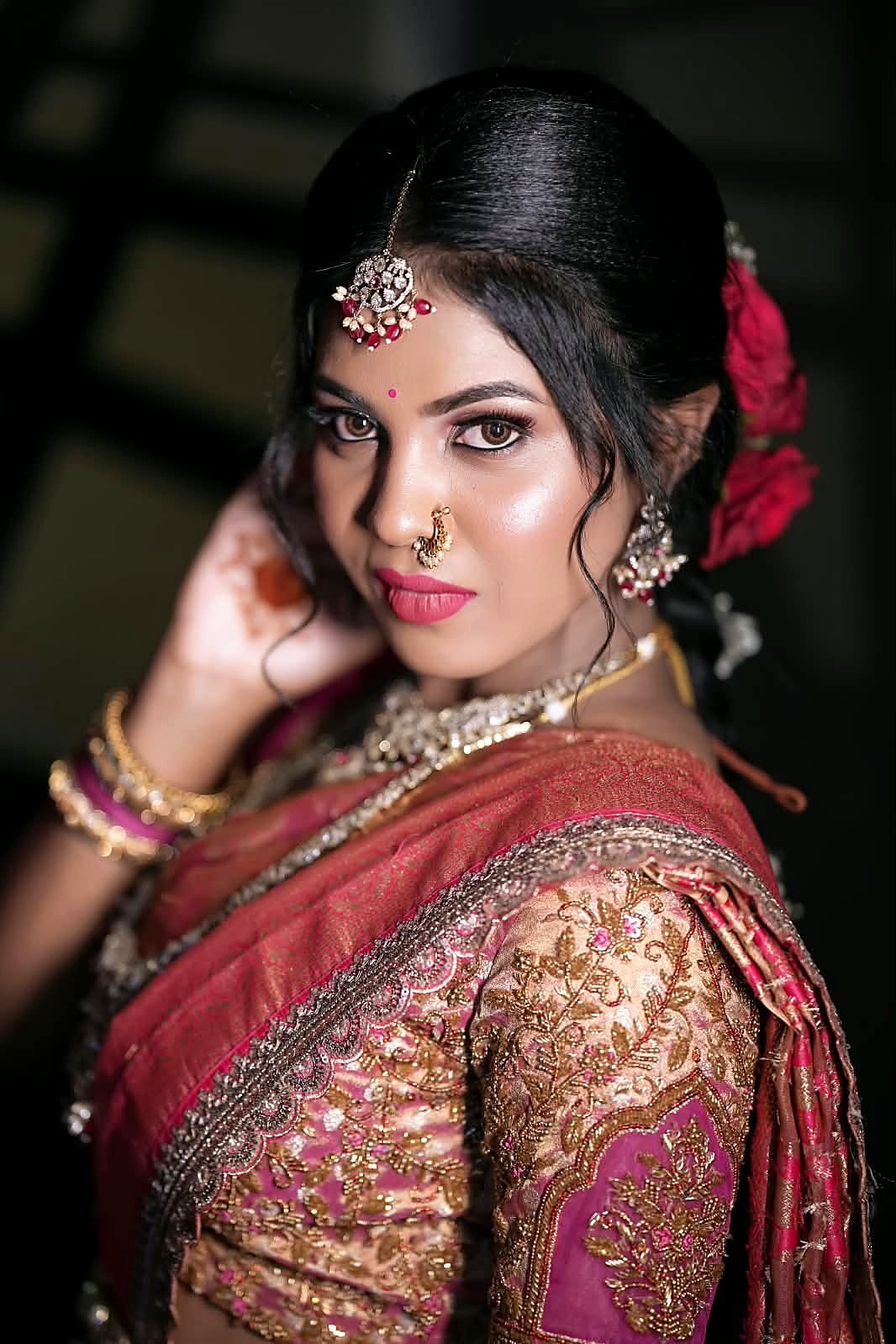 Bride looking over her shoulder with glowing skin, defined brown eyes and pink matte lips, wearing a maang tikka, nath and red roses in her hair
