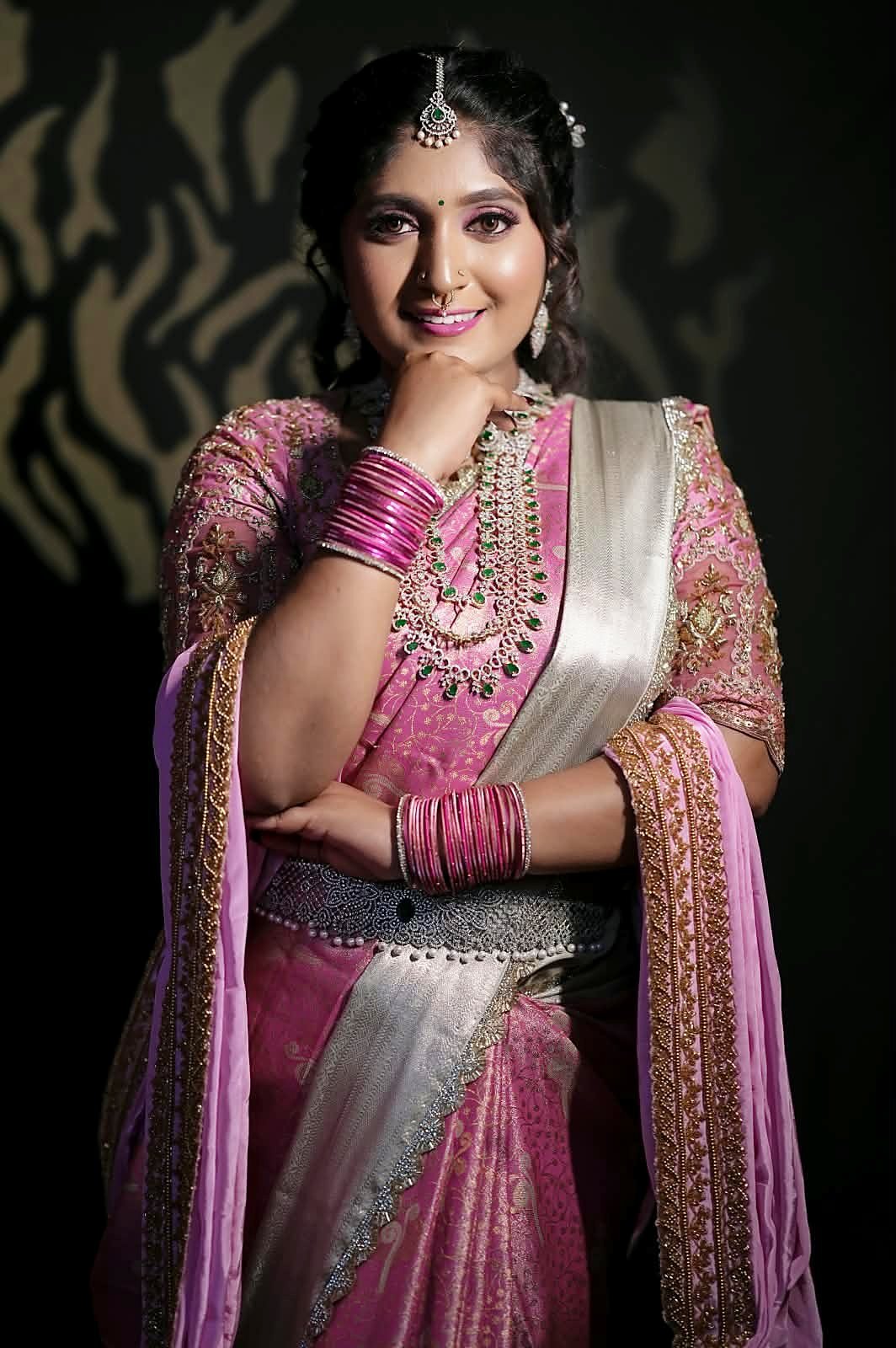 Smiling studio portrait of a woman in a pink silk saree with emerald-and-diamond jewellery and soft pink eye makeup