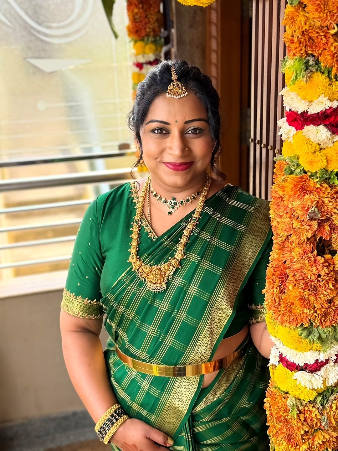 Smiling woman in a green and gold checked silk saree with temple jewellery, beside a marigold flower garland