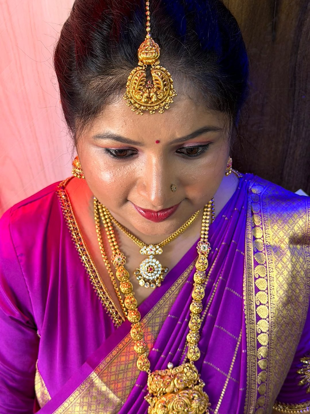Close-up of makeup with defined brows, warm shimmer lids and red lips, worn with a purple silk saree and gold temple jewellery